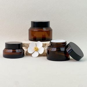 May include: Two amber glass jars with black lids and a white flower-shaped decorative element. One jar is closed, while the other is open with its black lid placed to the side, revealing a white inner seal. The jars have a wide, rounded base.