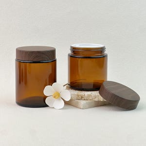 Könnte beinhalten: Zwei bernsteinfarbene Gläser mit Holzdeckeln. Ein Glas ist geschlossen, das andere ist offen. Eine weiße Blume mit gelbem Zentrum ist zwischen den Gläsern platziert. Die Gläser sind zylindrisch und stehen auf neutralem Hintergrund.