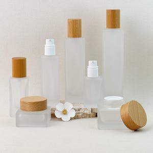 May include: A collection of frosted glass cosmetic containers featuring square-shaped bottles and jars with light wood grain caps. The set includes tall pump bottles, smaller pump bottles, and wide jars, some with lids removed to show the white inner rims.