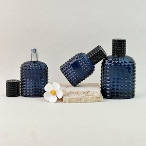 May include: Three navy blue perfume bottles with a geometric pattern. One bottle has a silver atomizer, another is lying on its side, and the third stands upright. A black cap and a white flower are also present.