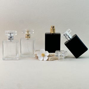 Pode incluir: Quatro frascos de perfume com corpos de vidro retangulares transparentes e borrifadores transparentes ou dourados. Um frasco é preto. Uma flor branca e uma tampa transparente também estão presentes. Os frascos estão dispostos em uma superfície neutra.