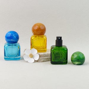 Close-up view of yellow, blue, and green colored glass perfume bottles with marbled caps, showing the spray nozzle and cap removed on neutral background.