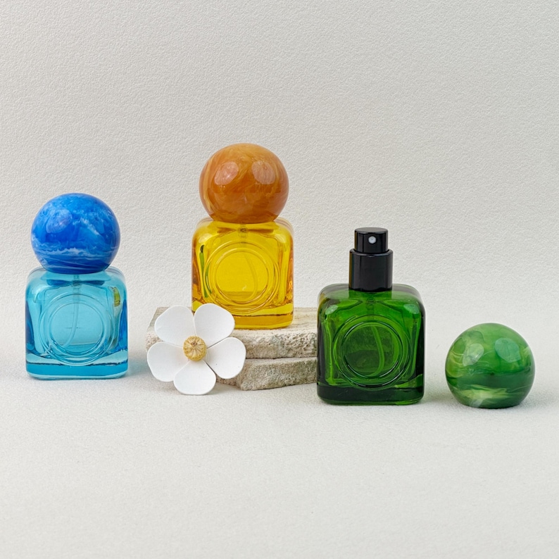 Close-up view of yellow, blue, and green colored glass perfume bottles with marbled caps, showing the spray nozzle and cap removed on neutral background.