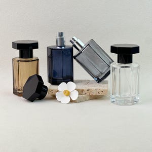 Op de afbeelding: Vier zeshoekige glazen parfumflesjes met zwarte doppen. Eén flesje is amberkleurig, één donkerblauw, één transparant en één grijs en gekanteld. Een witte bloemvormige decoratie met een geel hartje staat voor de flesjes.