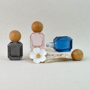 May include: Three small glass perfume bottles with octagonal shapes and round wooden caps. One bottle is dark gray, one is light pink, and one is blue with a rollerball applicator. A white five-petaled flower decoration sits in the center.