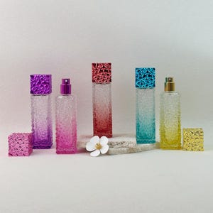 May include: An assortment of colorful, textured glass perfume bottles with matching square caps. The bottles are in shades of purple, pink, red, blue, and yellow. A white flower sits in front of the bottles.