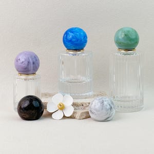 Pode incluir: Frascos de perfume de vidro transparente com tampas esféricas decorativas em mármore azul, verde e roxo. Também estão presentes tampas pretas e cinzentas, juntamente com uma flor branca. Os frascos têm detalhes dourados.