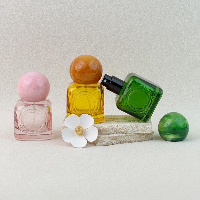 30ml pink, yellow, and green colored glass perfume bottles with marbled caps, one bottle tilted to show the spray nozzle on textured background.