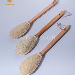 Puede incluir: Tres cepillos de espalda de madera con almohadillas de luffa. Los cepillos tienen mangos largos de color marrón claro y almohadillas de luffa de forma ovalada. Cada mango tiene un pequeño agujero con un lazo de fibra natural. La marca "Rafandra Craft" es visible.