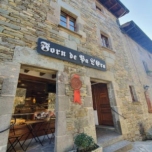 Peut inclure: Vue extérieure d'un bâtiment en pierre avec l'inscription "Forn de Pa L'Era" au-dessus de l'entrée. Un ruban rouge avec un sceau "50" est suspendu près de la porte. La citation "Oh! the places you'll go! - Dr. Seuss" est en bas.