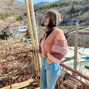 Könnte beinhalten: Eine Person trägt eine braune Strickmütze und eine rosa-lila Strickjacke. Die Strickjacke hat einen V-Ausschnitt und lange Ärmel. Sie trägt auch eine blaue Jeans und eine Sonnenbrille. Der Hintergrund zeigt eine ländliche Landschaft mit Bergen und blauem Himmel.