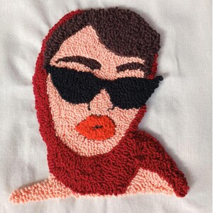 Op de afbeelding: Een punch needle kunstwerk met een gestileerd gezicht met een zwarte zonnebril en een rode sjaal. De lippen zijn rood en het haar is bruin. De achtergrond is wit.