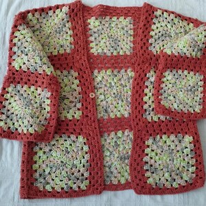 Può includere: Cardigan all'uncinetto fatto a mano con motivo a granny square. Il cardigan aperto è color corallo con quadrati multicolori in verde, giallo e grigio. Ha maniche lunghe e una chiusura con un solo bottone.