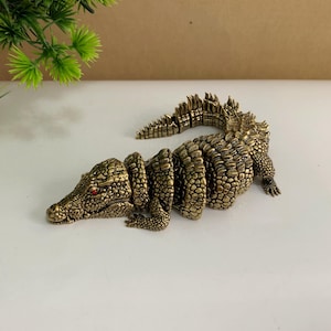 Könnte beinhalten: Eine detaillierte, antik anmutende Krokodilfigur aus Messing mit roten Augen. Der bewegliche Körper ist segmentiert und ermöglicht eine flexible Positionierung. Die Skulptur ist ein dekoratives Objekt mit aufwendigen Schuppen und einer strukturierten Oberfläche.