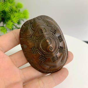 Op de afbeelding: Een bronzen schildpadschild-snijwerk. De schaal heeft een gedetailleerd patroon van geometrische vormen en lijnen. Het midden van de schaal heeft een Yin en Yang symbool. Het snijwerk wordt in een hand gehouden.