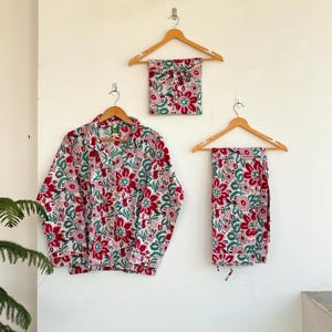 以下が含まれることがあります： 木製のハンガーに掛けられた、ボタンダウンシャツとパンツを含む花柄のパジャマセット。セットは、赤と緑の花の繰り返し模様の白い背景が特徴です。お揃いのスリープマスクも表示されています。