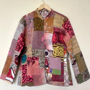May include: A patchwork jacket with a mandarin collar and long sleeves. The jacket is made of various fabric patches in shades of pink, brown, green, and gold. It has a button-up front and a small pocket on the front.