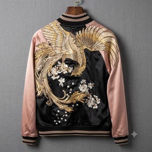 Puede incluir: Una chaqueta bomber de satén con un panel trasero negro que presenta un bordado de fénix dorado y detalles florales blancos. Las mangas y los paneles frontales son de color rosa claro, con cuello, puños y dobladillo acanalados a rayas.