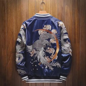 Puede incluir: Una chaqueta bomber de satén azul marino con un intrincado bordado plateado de un fénix y una luna creciente dorada en la espalda, motivos de dragón en las mangas, cuello, puños y dobladillo acanalados con rayas blancas en una percha.