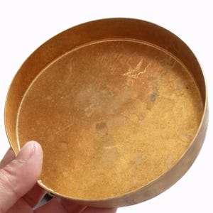 May include: A round, brass-colored bowl with a slightly raised edge. The bowl has a smooth, reflective surface with some minor imperfections. The bowl is held by a hand against a white background. The bowl is likely used for decorative or functional purposes.