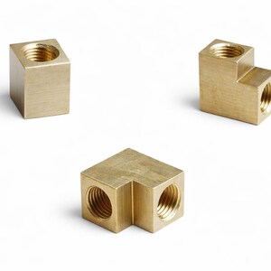 May include: Three brass pipe fittings are shown against a white backdrop. The fittings include a cube, an L-shaped piece, and a corner piece, all with threaded openings. These are likely used in plumbing or engineering.