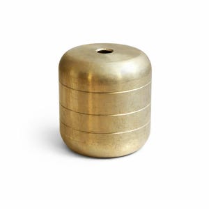 Puede incluir: Un recipiente cilíndrico de latón con la parte superior e inferior redondeadas. El recipiente tiene tres ranuras horizontales y un pequeño agujero en el centro de la parte superior. El objeto es de metal brillante.