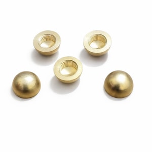 Puede incluir: Cinco componentes de hardware de latón en forma de media esfera. Cuatro tienen un interior roscado y uno es una cúpula sólida. Los componentes son de color dorado cálido y se encuentran sobre un fondo blanco.