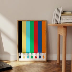 Pode incluir: Uma impressão artística Bauhaus emoldurada com listras verticais em preto, amarelo, turquesa, vermelho, azul e laranja. A palavra "BAUHAUS" é impressa em letras coloridas na parte inferior sobre um fundo branco. A impressão está em uma moldura de madeira.