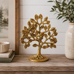 May include: A gold-colored metal tree sculpture featuring a textured trunk, exposed roots, and numerous detailed leaves. The decorative object stands on a circular base and displays a symmetrical branch structure with a polished metallic finish.