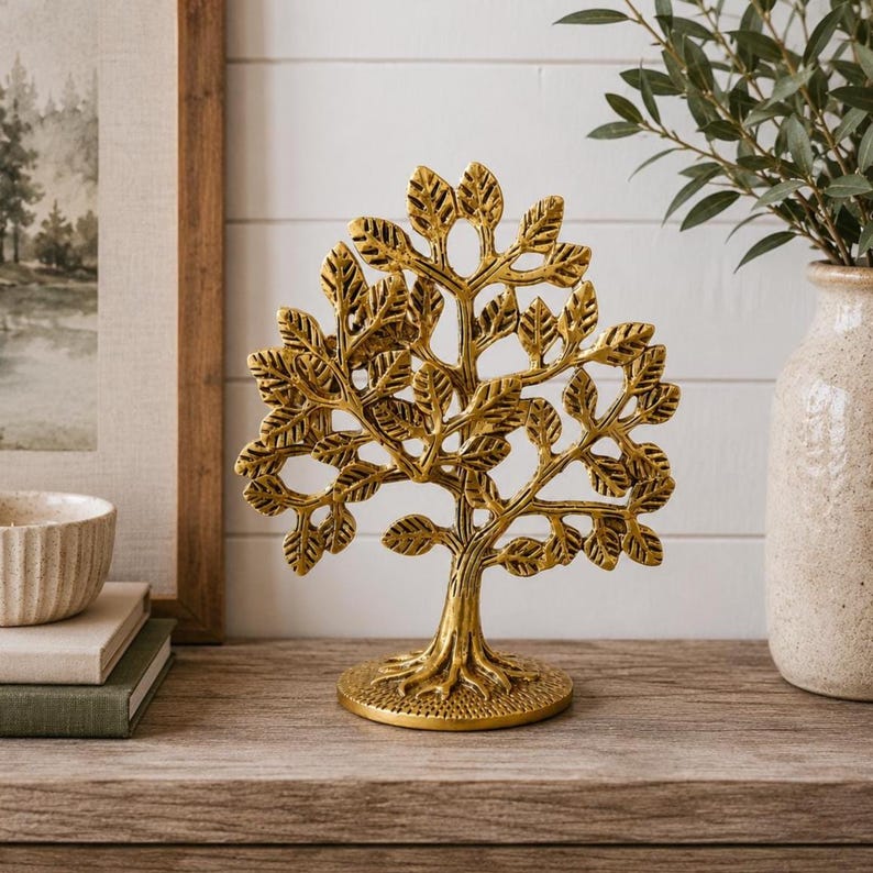 May include: A gold-colored metal tree sculpture featuring a textured trunk, exposed roots, and numerous detailed leaves. The decorative object stands on a circular base and displays a symmetrical branch structure with a polished metallic finish.