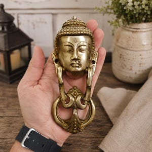 Pode incluir: Um aldravão de porta em latão polido com uma cabeça de Buda esculpida, olhos fechados e um ushnisha texturizado. O design inclui um anel oscilante decorativo e ornamentado fixado na base da cabeça com detalhes metálicos intrincados.