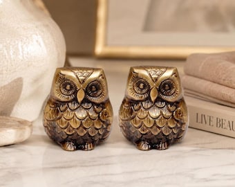 May include: Two bronze-colored owl figurines with detailed feather and eye designs. The owls are sitting side-by-side on a marble surface. The figurines have a vintage or antique aesthetic, suitable for home decor.