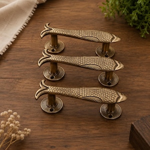 Op de afbeelding: Drie bronzen deurgrepen met een visontwerp. Elke handgreep heeft een visvormig lichaam met schubben en een vinvormig uiteinde. De handgrepen zijn gemonteerd op ronde bases, wat een decoratieve set creëert.