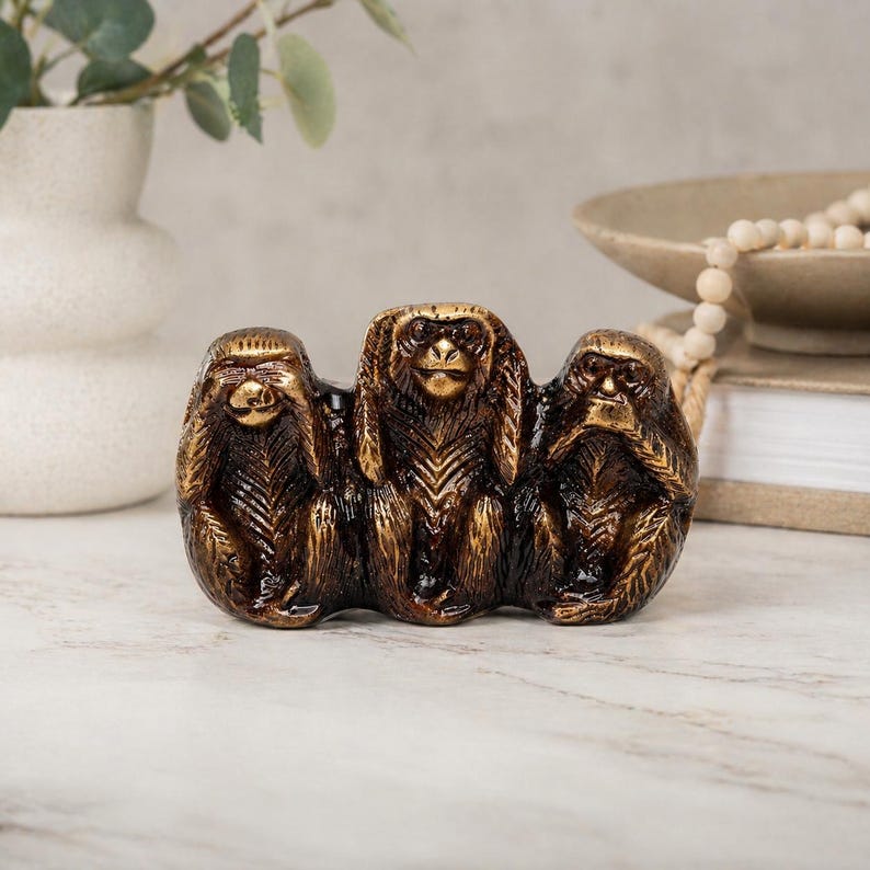 May include: A bronze-colored sculpture of the three wise monkeys: See no evil, hear no evil, speak no evil. The monkeys are seated together, with their hands covering their eyes, ears, and mouth. The sculpture has a textured finish.