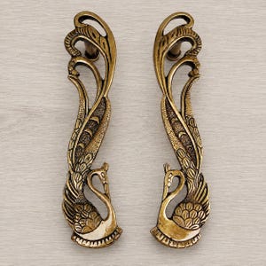 Puede incluir: Un par de manijas de puerta verticales de latón con un intrincado diseño de pavo real. Cada manija muestra un ave estilizada con plumaje detallado, cuello curvado y una cola larga y fluida que se extiende hacia arriba, en tono dorado.