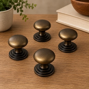 May include: A set of four round cabinet knobs with a brushed antique brass finish. Each knob features a smooth, domed top and a tiered, circular base that connects to the stem, providing a consistent look for furniture or cabinetry hardware.