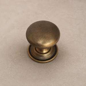 May include: A round cabinet knob with a brushed antique brass finish. The knob features a smooth, domed top surface and a circular base plate with a stepped design, showcasing a weathered metallic texture throughout the entire hardware piece.