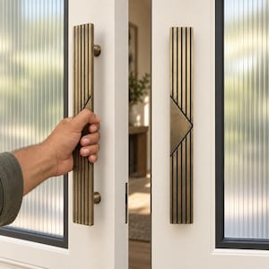 May include: A pair of long, vertical door handles made of brushed brass with a series of parallel black grooves running the length of the metal. Each handle features a central triangular cutout design and is mounted on a white door frame.