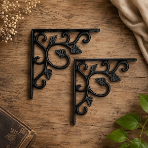 May include: Two black metal shelf brackets featuring an L-shaped design with decorative scrollwork and leaf motifs. Each bracket has pre-drilled holes for mounting and a smooth, dark finish, showcasing intricate swirling patterns and foliage details.