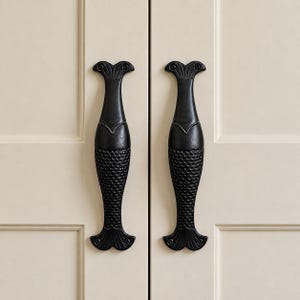 May include: Two vertical black cabinet handles shaped like fish, mounted on a cream-coloured double door. Each handle features a detailed scale pattern on the lower body, a smooth midsection, and a flared tail design at the top and bottom ends.