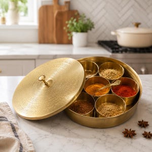 May include: A round brass spice box with an etched lid and seven internal containers filled with various colorful ground spices. The container features a central knob on the lid and a metallic finish, providing a storage solution for kitchen spices.