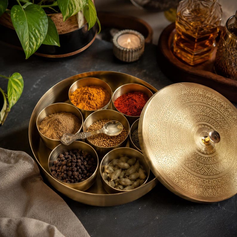 May include: A round brass spice container with a removable lid featuring an intricate etched pattern and a central knob. The container holds seven small, removable brass bowls filled with various colorful spices and a small ornate metal spoon.