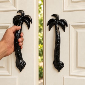 Puede incluir: Un par de manijas de puerta negras con forma de palmeras, con troncos texturizados y frondas curvas en la parte superior. Las manijas están montadas verticalmente en puertas dobles blancas con paneles y presentan un diseño detallado.
