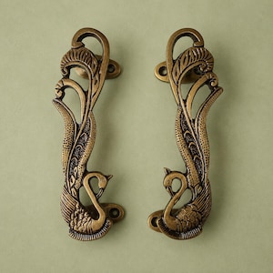 Può includere: Una coppia di maniglie per porta in ottone antico con un design dettagliato a forma di pavone. Ogni maniglia presenta un collo curvo e una lunga coda fluida con complesse texture di piume. Il metallo ha una finitura scura e invecchiata.