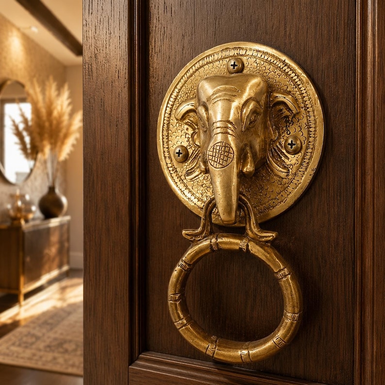 May include: A brass elephant-shaped door knocker with a circular handle. The knocker is mounted on a dark brown wooden door. The elephant head is detailed with a textured surface and is attached to a round plate with four screws.
