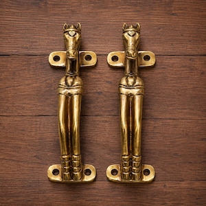 May include: Two brass door handles with a horse head design. The handles are gold-colored and feature a detailed horse head at the top, with a decorative element on the forehead. The handles are mounted on a wooden surface.