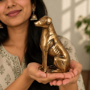 May include: A bronze-colored dog statue in a seated position. The sculpture has detailed features, including etched lines that define the dog's form. The statue is held in open hands, showcasing its metallic sheen and craftsmanship. A decorative item.