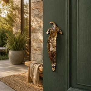 May include: A gold-colored, leaf-shaped door handle is mounted on a dark green door. The handle has intricate detailing and is attached with two small, dark-colored screws. The door is part of a house exterior, with a stone wall and greenery visible in the background.
