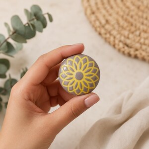 Puede incluir: Un pomo de cerámica circular con un acabado gris liso y brillante, decorado con un patrón floral o de mandala amarillo vibrante y estilizado en la parte frontal. El diseño presenta un círculo central rodeado de pétalos radiantes e interconectados.