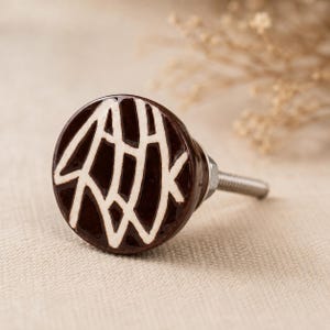May include: A round, dark brown ceramic cabinet knob with a white abstract design. The knob has a shiny finish and a silver metal screw for attachment. This decorative hardware is suitable for drawers and cabinets.